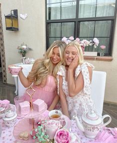 two women are sitting at a table with tea cups and plates on it, posing for the camera