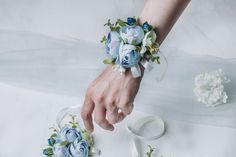 a bride's bracelet with blue flowers and ribbons on her wrist, next to the wedding dress