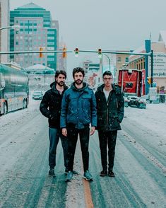 three men standing in the middle of a snowy street with traffic lights and buildings behind them