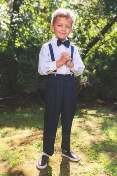 a young boy wearing a bow tie and suspenders standing in the grass with his hands folded