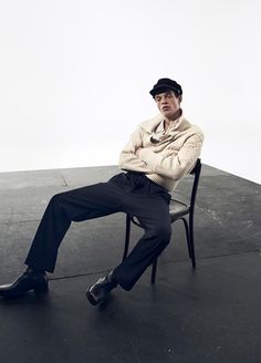 a man is sitting on a chair with his legs crossed, wearing a hat and sweater