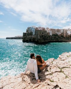 two people sitting on the edge of a cliff overlooking the ocean and buildings in the background