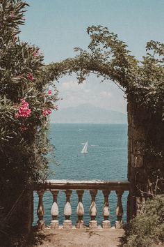 an archway leading to the ocean with a sailboat in the distance and pink flowers on either side