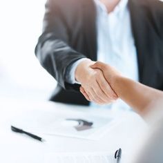 two people shaking hands at a table with papers and pens on it, in front of them