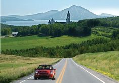 a red car driving down the road in front of a church with a mountain view