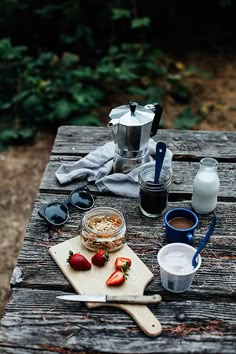 a picnic table with coffee, milk and strawberries on it in front of the woods