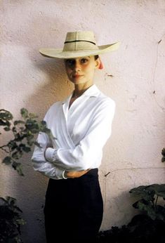 a woman wearing a hat and holding a flower in her hand while standing next to a wall