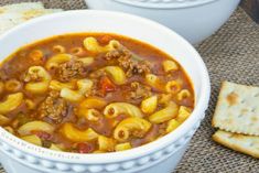 two bowls filled with pasta and meat soup next to crackers on a tablecloth