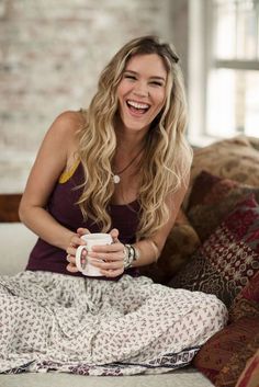 a woman sitting on a couch holding a coffee cup and smiling at the camera with her legs crossed