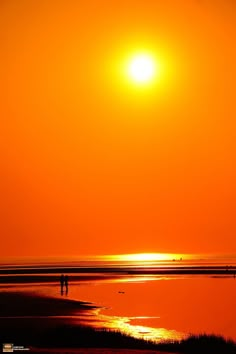 two people are walking on the beach as the sun goes down in the sky over the water