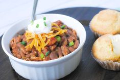 a white bowl filled with chili and cheese next to muffins