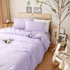 a bed with purple sheets and pillows in a room next to a small tree on the floor