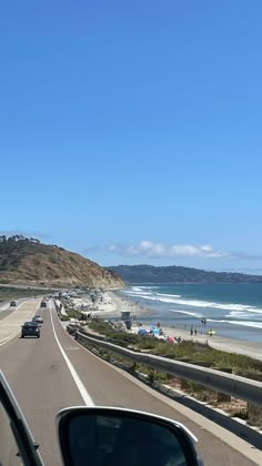 cars driving down the road next to the beach and ocean on a sunny day with blue skies