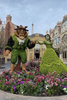 a couple of people that are standing in the grass near some flowers and bushes with buildings in the background