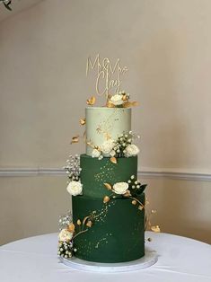 a three tiered green wedding cake with white flowers and gold lettering on top, sitting on a round table