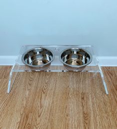 two stainless steel bowls on clear acrylic stand with wood floor and wall in background