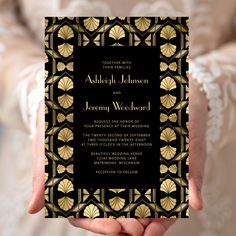 a person holding up a black and gold wedding card with an art deco design on it