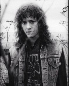 a man with long hair wearing a jean jacket and standing in front of some trees