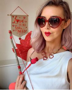 a woman with pink hair and heart shaped sunglasses holding a stick that says love is all you need