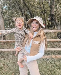 a woman holding a child in front of a fence with trees and grass behind her