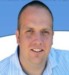 a man in a blue shirt and tie smiling at the camera with his name on it