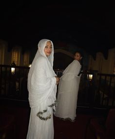 two women dressed in white standing next to each other on a balcony at night time