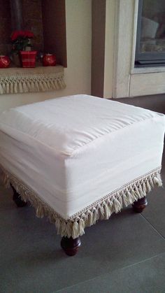 a white ottoman with tassels on it sitting in front of a window next to a potted plant