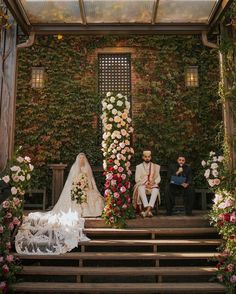 a bride and groom are sitting on the steps in front of a floral covered wall