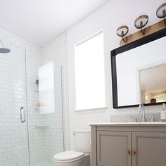 a white toilet sitting next to a walk in shower under a bathroom mirror on top of a wooden cabinet