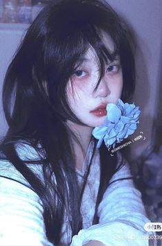 a woman with long black hair holding a blue flower in her mouth and looking at the camera