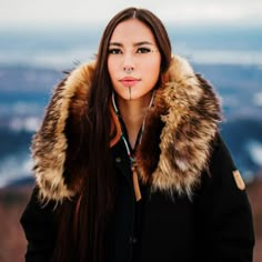 a woman with long brown hair wearing a black jacket and fur collar standing on top of a mountain