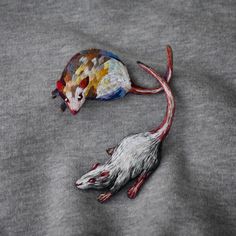 two rat sculptures are laying on top of a gray shirt, one is painted with multicolored paint