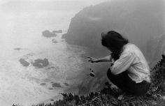 a woman kneeling down on top of a hill next to the ocean and holding something in her hand