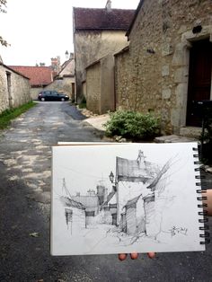 a person holding up a drawing in front of an alley way with cars parked on the side