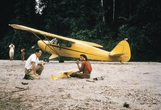 two people are sitting on the ground next to a small plane and another person is taking pictures