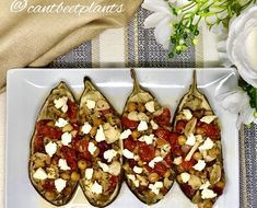 three stuffed eggplant with tomatoes and cheese on a white plate next to flowers