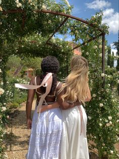 two women walking down a path under a pergolia covered arbor with white flowers