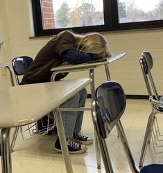 a person sitting at a desk with their head on the back of another chair, in front of a window