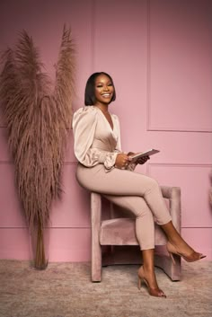 a woman sitting on a chair in front of a pink wall and holding a tablet