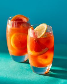 two glasses filled with orange juice and lemon slices on a blue tablecloth next to each other