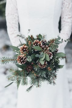 a bride holding a bouquet of pine cones and greenery in the snow with her hands