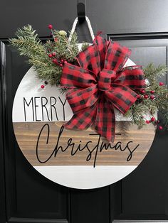 a merry christmas sign hanging on the front door with pine branches and red plaid bow