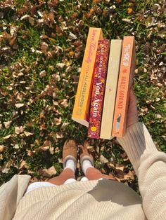 a person is holding three books in their hands while sitting on the grass with leaves