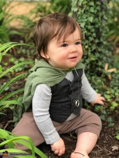 a baby sitting on the ground wearing a green vest and smiling at the camera with plants in the background