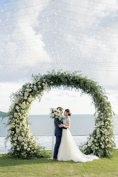 a bride and groom standing under an arch with white flowers on the grass by the water