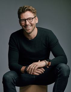 a man with glasses sitting on top of a wooden stool smiling at the camera while wearing a black sweater and jeans