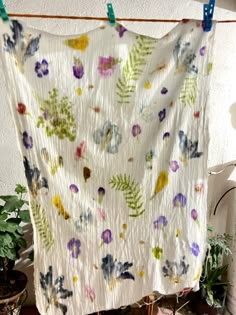 a white quilt hanging from a clothes line with flowers and leaves on it in front of a potted plant