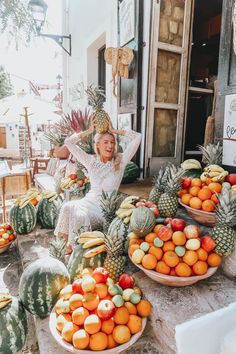 a woman sitting in front of a display of fruit on a table with pineapples, oranges and melons