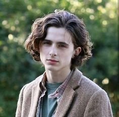 a young man with curly hair wearing a brown jacket and green shirt standing in front of trees