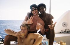 two men and a woman sitting on a boat with drinks in front of them, making the peace sign